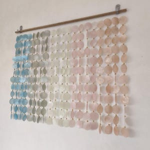 May include: A wall hanging featuring multiple vertical strands of circular, translucent shell discs suspended from a horizontal wooden rod. The discs transition in colour from light blue and grey on the left to white, pink, and peach on the right.