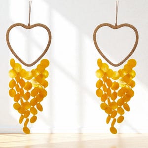 Peut inclure: Deux décorations murales en forme de cœur. Chacune a un cadre marron tressé et une cascade d'accents de coquillages jaunes de forme ovale. Les décorations sont suspendues à une corde marron, créant une esthétique décorative et naturelle.