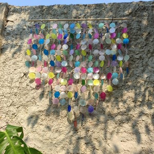 May include: A wall hanging composed of multiple vertical strands of circular, translucent, multi-colored shell discs suspended from a horizontal wooden rod. The discs feature a variety of hues including pink, blue, yellow, purple, and white.