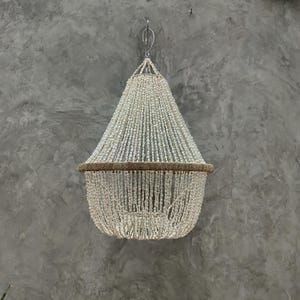 May include: A chandelier made of small, white, shell-like beads, suspended from a metal hook. The chandelier has a conical shape, with a brown band around the middle. The background is a textured gray wall.