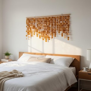 Könnte beinhalten: Ein Wandbehang bestehend aus zahlreichen kleinen, kreisförmigen, bernsteinfarbenen Holzscheiben, die an einer horizontalen Holzstange hängen. Die Scheiben sind in vertikalen Strängen unterschiedlicher Länge angeordnet und erzeugen einen kaskadierenden Effekt.