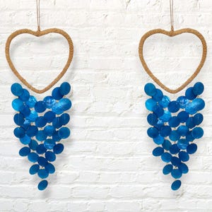 May include: Two heart-shaped wall hangings. Each has a woven rope heart frame and a cascade of blue, round, shell-like pieces. The hangings are suspended by a thin, brown string against a white brick wall.