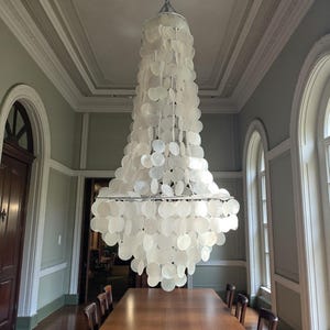 Puede incluir: Una lámpara de araña grande y elegante con un diseño escalonado, compuesta por numerosos elementos blancos en forma de concha. Cuelga sobre una larga mesa de comedor de madera en una habitación con techos altos y ventanas arqueadas.