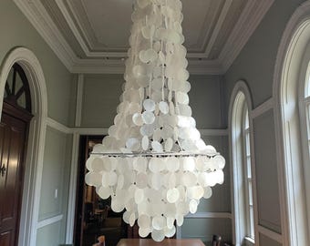 May include: A large, elegant chandelier with a tiered design, composed of numerous white, circular shell-like elements. The fixture hangs above a long, wooden dining table in a room with high ceilings and arched windows.