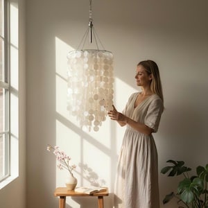 May include: A decorative chandelier with a silver metal frame and cascading, round, off-white shell accents. The light fixture hangs from a chain. The image shows a woman in a beige dress standing near the chandelier.
