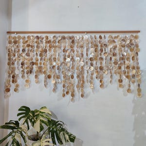 May include: A wall hanging consisting of multiple vertical strands of circular, iridescent capiz shell discs suspended from a horizontal wooden rod. The translucent, off-white and tan shells vary in tone and are arranged in a cascading pattern.