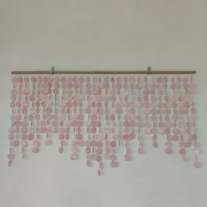 May include: A wall hanging featuring multiple vertical strands of pale pink circular discs suspended from a horizontal wooden rod. The light pink discs are arranged in varying lengths, creating a layered, cascading effect across the wall.