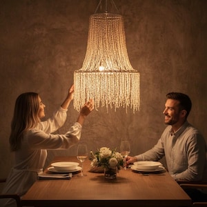 Puede incluir: Una lámpara de araña de cuentas cuelga sobre una mesa de comedor de madera preparada para una comida. La lámpara de araña está hecha de hebras de cuentas, y la mesa está puesta con platos, vasos y un centro de mesa floral. Una pareja está sentada a la mesa.