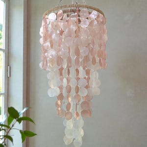 Può includere: Un lampadario decorativo con un design a strati, caratterizzato da numerosi pezzi rotondi e iridescenti a forma di conchiglia nei toni del rosa e del bianco. Il lampadario è sospeso a un anello avvolto in corda naturale.