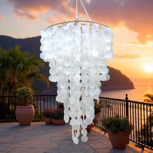 May include: A white capiz shell chandelier with a tiered design, suspended from a chain. The chandelier features numerous round, translucent shells, creating a cascading effect. The fixture is designed for indoor or outdoor use.