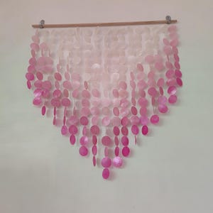 Peut inclure: Décoration murale composée de rangées de disques roses et blancs en forme de coquillages. Le motif est disposé en cascade, le rose s'intensifiant vers le bas. Suspendu à une baguette en bois, il crée un effet décoratif.