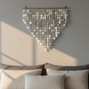Könnte beinhalten: Wandbehang aus kleinen, runden, irisierenden Muschelstücken. Das Kunstwerk hängt an einer natürlichen Holzstange und bildet eine kaskadenartige, dreieckige Form. Es befindet sich über einem Bett mit neutralfarbenen Kissen und Bettwäsche.