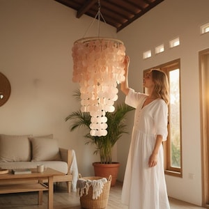 May include: A large, tiered chandelier with numerous, round, light pink shell-like elements hangs from the ceiling. The fixture is suspended in a well-lit room with a woman in a white dress standing nearby.