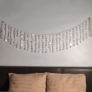 May include: A decorative wall hanging composed of strings of white, shell-shaped discs. The discs are strung on a thin, silver wire, forming a curved, cascading pattern. The wall hanging is above a bed with tan pillows.