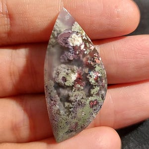 May include: A teardrop-shaped, translucent agate cabochon with a unique pattern of green, purple, and red dendrites. The stone has a natural, organic look and would be perfect for jewelry making.