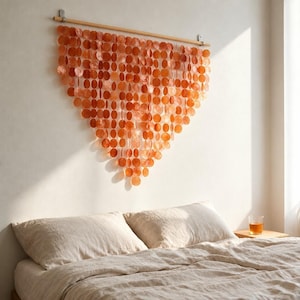 May include: A wall hanging composed of numerous translucent, circular orange discs suspended from a horizontal wooden rod. The discs are arranged in vertical strands that form a V-shaped pattern, creating a layered, semi-transparent display.