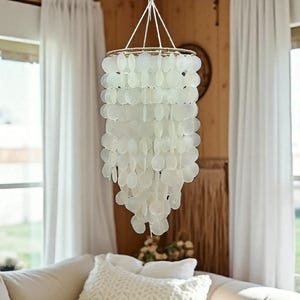 May include: A white capiz shell chandelier hangs from the ceiling. The chandelier has a circular frame and is decorated with many, overlapping, oval-shaped capiz shells. The light fixture is suspended by white cords.