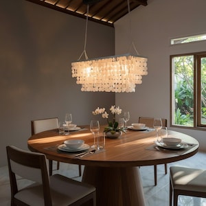 May include: A rectangular chandelier featuring multiple tiers of translucent, circular white shell discs suspended from a metal frame by thin chains, emitting a soft glow above a round wooden dining table set with white bowls and glassware.