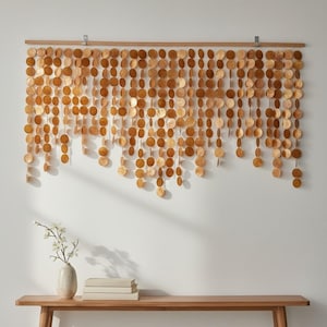 May include: A wall hanging consisting of numerous vertical strands of circular, golden-brown discs suspended from a horizontal wooden rod. The discs vary slightly in size and tone, creating a layered, textured appearance across the wall surface.