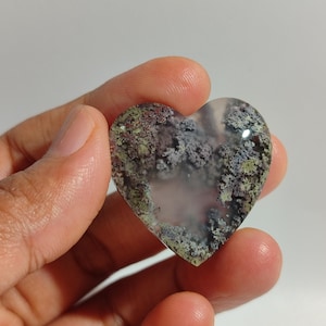 Puede incluir: Un cabujón de ágata en forma de corazón con un centro blanco translúcido y un patrón dendrítico de inclusiones verdes, moradas y marrones.