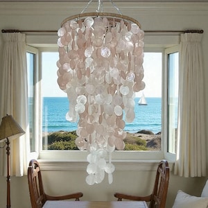 Op de afbeelding: Een decoratieve kroonluchter met een houten frame en ronde, schelpvormige elementen in roze en wit. De lamp hangt voor een raam met uitzicht op zee, inclusief een zeilboot. De kamer heeft een kustthema.
