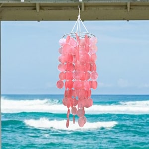 Puede incluir: Un carillón de conchas rosa colgando de un techo blanco. El carillón está hecho de múltiples capas de conchas rosas que están enhebradas juntas. El carillón está colgando frente a un océano azul con olas blancas.