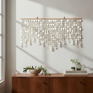 May include: A wall hanging composed of numerous small, circular, iridescent white shell discs suspended from a horizontal wooden rod. The discs are arranged in multiple vertical strands of varying lengths, creating a layered, textured appearance.