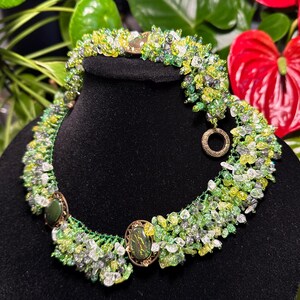 Puede incluir: Un collar de cuentas verdes, blancas y amarillas con dos colgantes grandes de forma ovalada, verdes y dorados. El collar se muestra en un maniquí de terciopelo negro.