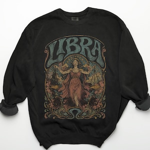 May include: A black crewneck sweatshirt featuring a graphic print of a figure holding scales. The design includes the text LIBRA in a stylised font, surrounded by floral motifs and clouds in muted tones of blue, pink, and gold on the front centre.