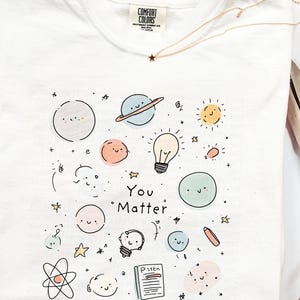 May include: White t-shirt featuring a colourful illustration of planets, stars, light bulbs, and the words "You Matter." The shirt has a crew neck and a small tag at the neckline. A gold necklace with a star pendant is also visible.