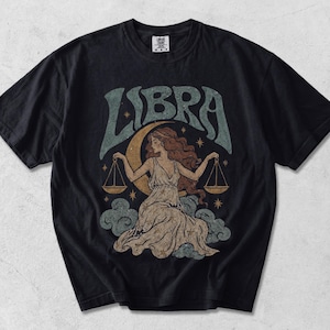 Puede incluir: Una camiseta negra de manga corta con un estampado gráfico de una persona con pelo largo sosteniendo una balanza, frente a una luna creciente y nubes. La palabra LIBRA está impresa en letras grandes, estilizadas y de color verde azulado.