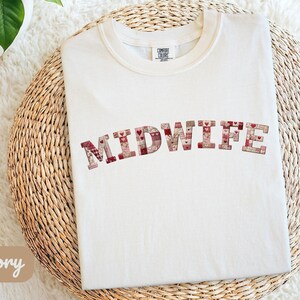 Peut inclure: T-shirt ivoire avec le mot "MIDWIFE" dans un motif patchwork de cœurs et de motifs rouges, roses et beiges. Le t-shirt est plié sur une surface circulaire tissée.