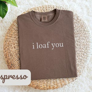 Può includere: T-shirt color espresso con la scritta bianca "i loaf you". La maglietta è piegata su un tappetino rotondo in vimini. La parola "espresso" è scritta su un'etichetta bianca. Una pianta in vaso è visibile sullo sfondo.
