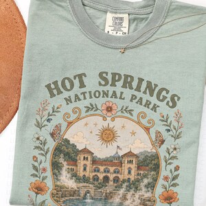 May include: A sage green short-sleeve t-shirt featuring a graphic print that reads HOT SPRINGS NATIONAL PARK. The design includes a central illustration of a building, a sun, butterflies, and floral borders in muted orange, brown, and cream tones.