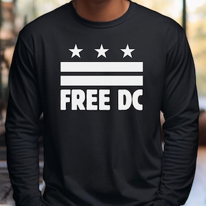 Peut inclure: T-shirt noir à manches longues avec un motif blanc. Le motif comprend trois étoiles au-dessus de deux lignes horizontales et l'inscription "FREE DC". Le t-shirt est en matière souple.
