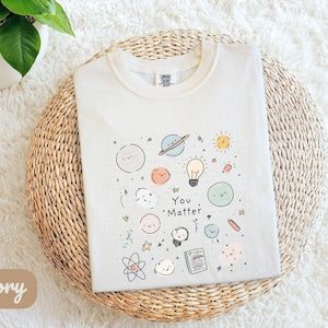May include: Ivory t-shirt featuring a playful design of planets, stars, and smiling faces, with the words "You Matter" printed below. The shirt is folded on a woven, circular mat.