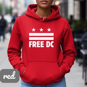 Puede incluir: Una sudadera con capucha roja con un gráfico blanco que incluye tres estrellas, dos líneas horizontales y las palabras "FREE DC". La sudadera tiene un bolsillo delantero y una capucha con cordón.