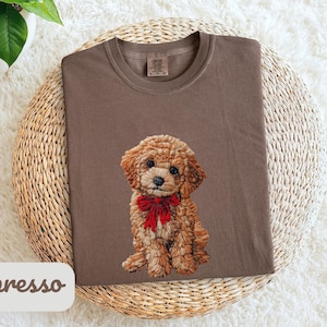 May include: Espresso-colored t-shirt featuring an embroidered poodle with a red bow. The dog is detailed with brown and tan thread. The shirt is folded on a woven mat, with a white label that says "espresso".