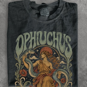 May include: A charcoal grey t-shirt featuring a graphic print with the text OPHIUCHUS in large, curved lettering. The illustration depicts a figure with long hair holding a large snake, surrounded by stars, clouds, and flowers in muted tones.