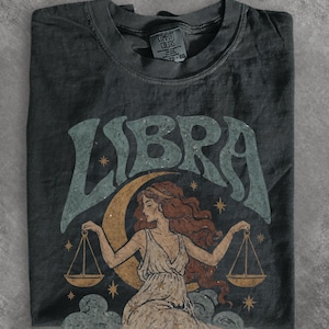 Könnte beinhalten: Ein anthrazitfarbenes T-Shirt mit einem Grafikdruck des Wortes LIBRA in großen, abgenutzten blaugrünen Buchstaben. Unter dem Text befindet sich die Illustration einer Figur mit langem Haar, die Waagschalen hält, vor einem Halbmond, Sternen und Wolken.