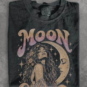 Könnte beinhalten: Ein anthrazitfarbenes T-Shirt mit dem Wort MOON in großen, farbverlaufenden Buchstaben. Unter dem Text befindet sich die Illustration einer Person mit langem, welligem Haar, das mit Blumen geschmückt ist, neben einer großen Mondsichel.