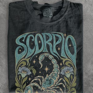Könnte beinhalten: Ein anthrazitfarbenes Kurzarm-T-Shirt mit einem großen Grafikdruck eines Skorpions, der unter dem Wort SCORPIO in einer stilisierten Schrift zentriert ist. Das Design enthält florale Elemente, kleine Sterne und eine verwaschene Farbpalette in Petrol und Olivgrün.