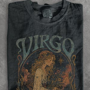 May include: A charcoal grey short-sleeve t-shirt featuring a graphic print of a woman with long flowing hair adorned with flowers. The text VIRGO is printed in a large, stylised font above the illustration, which includes stars and floral elements.