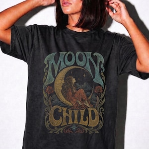 Könnte beinhalten: Ein anthrazitfarbenes Kurzarm-T-Shirt mit einem Grafikdruck einer Person, die auf einer Mondsichel sitzt. Der Text MOON CHILD ist in großen, stilisierten Buchstaben über und unter der Illustration gedruckt, umgeben von Blumen- und Sternmotiven.