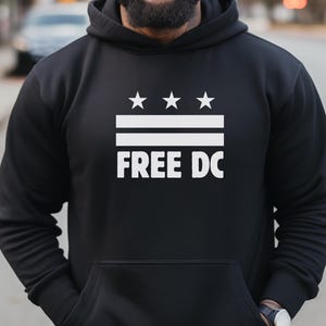May include: Black hoodie with a white graphic. The graphic includes three stars above two horizontal lines, with the words "FREE DC" below. The hoodie has a front pocket and a drawstring hood.