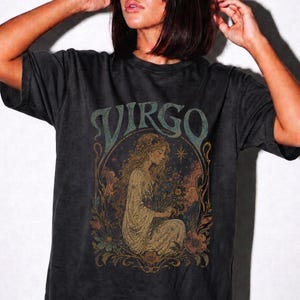 May include: A charcoal grey short-sleeve t-shirt featuring a graphic print of a person with long hair surrounded by flowers and foliage. The word VIRGO is printed in large, stylised letters above the illustration in a muted teal colour.