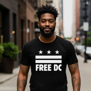 May include: Black t-shirt with a white graphic design featuring three stars, two horizontal lines, and the text "FREE DC". The shirt is worn by a person standing on a city street.