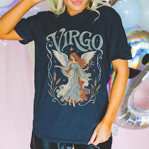 May include: A black short-sleeved t-shirt featuring a graphic print of a winged figure with long hair, surrounded by wheat stalks and stars. The word VIRGO is printed in large, stylised, light-coloured lettering above the central figure.