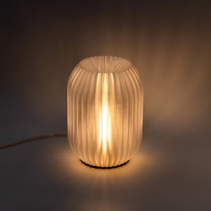 Puede incluir: Una lámpara de mesa blanca con una pantalla plisada translúcida. La lámpara está encendida y proyecta un brillo cálido.