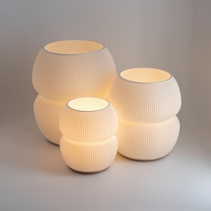 Op de afbeelding: Drie witte, ronde, geplooide lampenkappen in verschillende maten. De lampenkappen zijn van binnenuit verlicht en geven een warm licht.